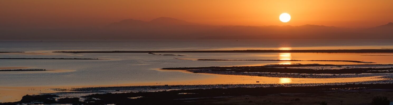 Sun setting over the bay, the sun is reflected in the water. The sky is a rich orange and reflected in the water.