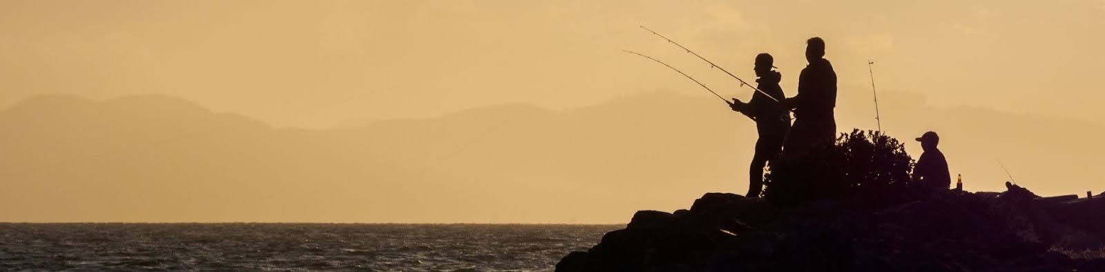 The people are in silhouette - two are standing on a rocky shoreline and fishing and a child is seated watching them. The sky is smoky beige with the outline of hills visible in the background. A small strip of Bay water is visible and is a dark grey-blue.