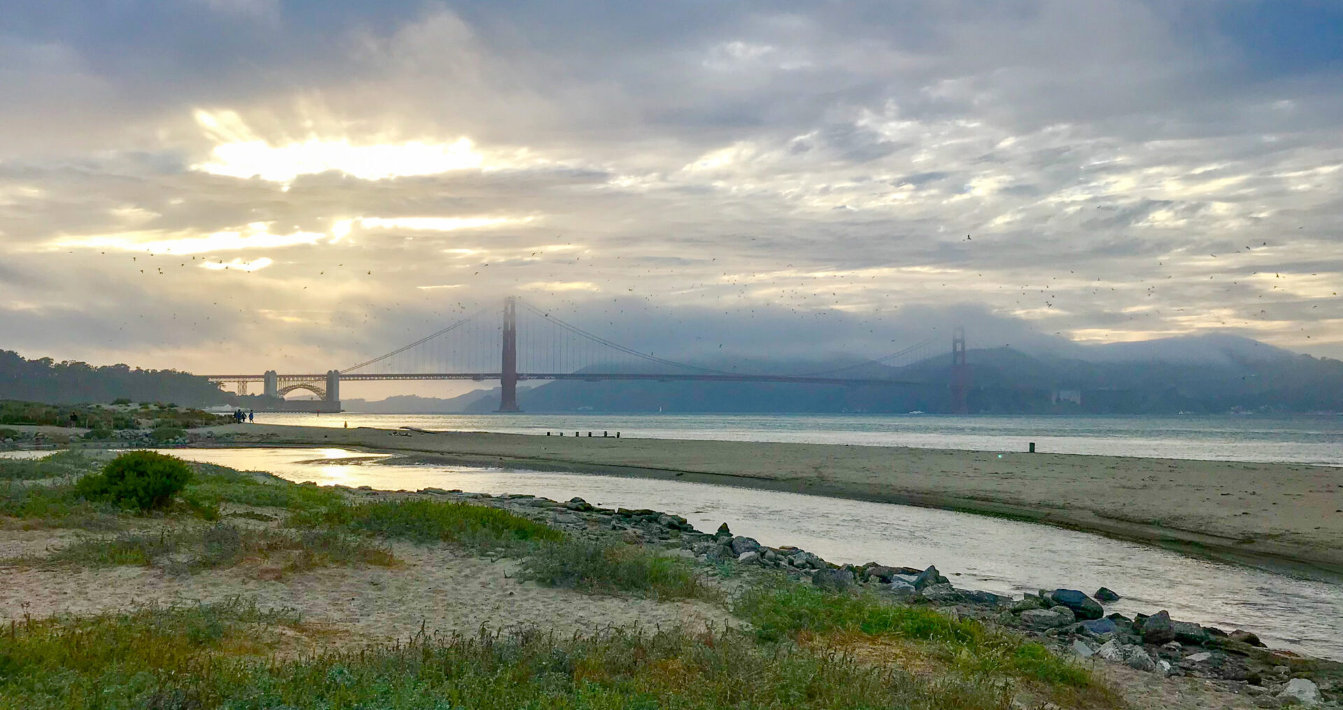 Light peeks through clouds over the Golden Gate Bridge, with water, a sandy beach, and vegetation in the foreground.