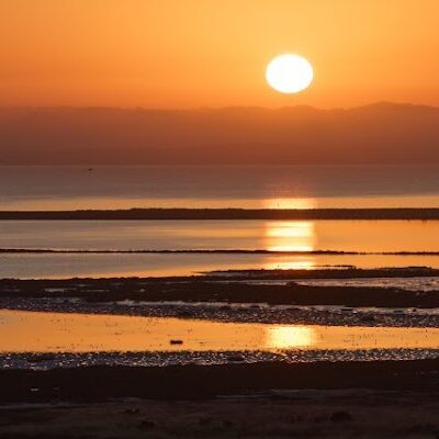 Sun setting over the bay, the sun is reflected in the water. The sky is a rich orange and reflected in the water.