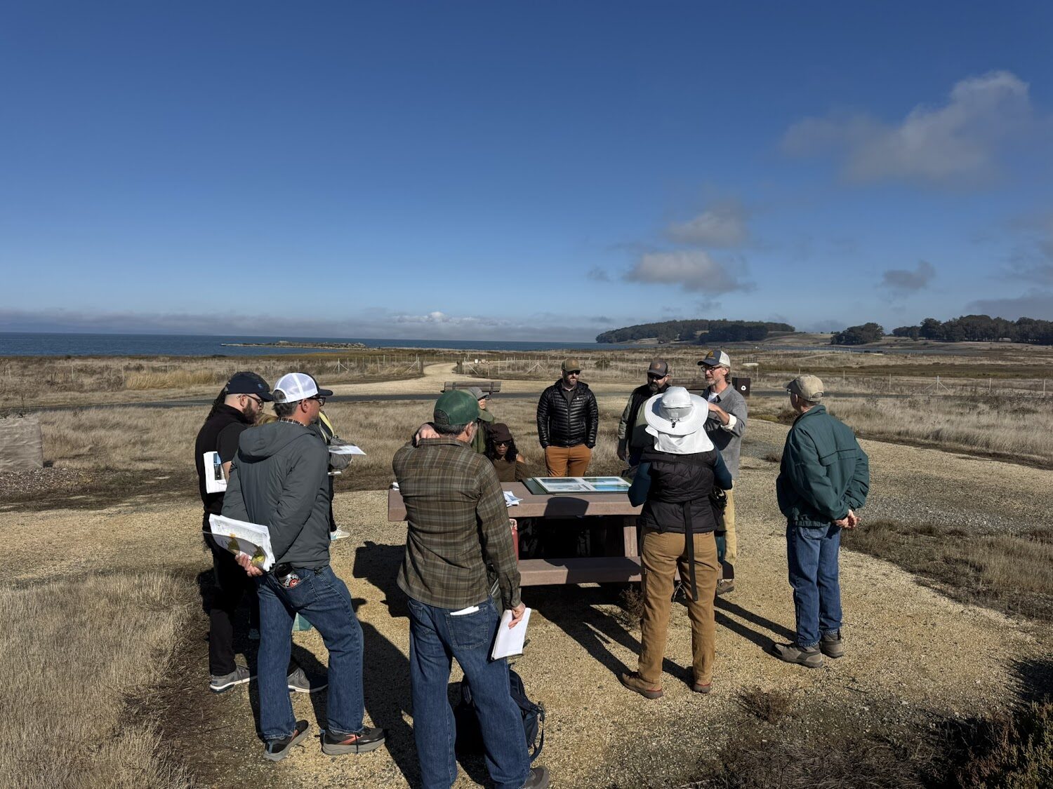 A group of people stand around a bunch with a marsh and trail behind them.